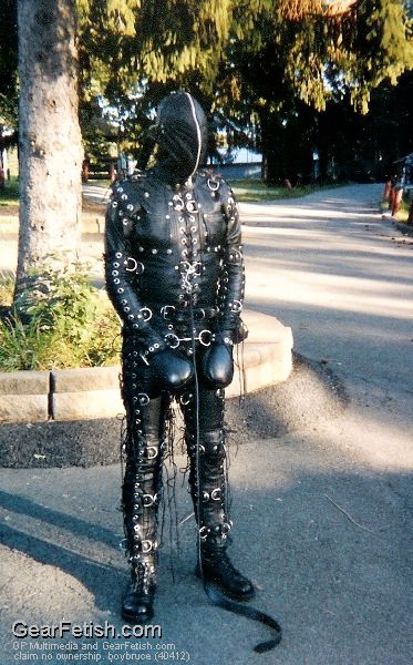A person, unable to see, is standing near a roundabout with a large tree in it. They are in head to toe leather, wearing a bondage suit, hood and fist mitts.
