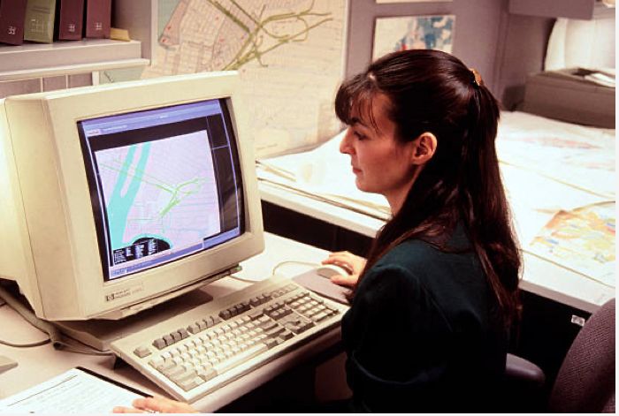 A 1990s office worker with bangs and shoulder pads edits something on a beige desktop computer