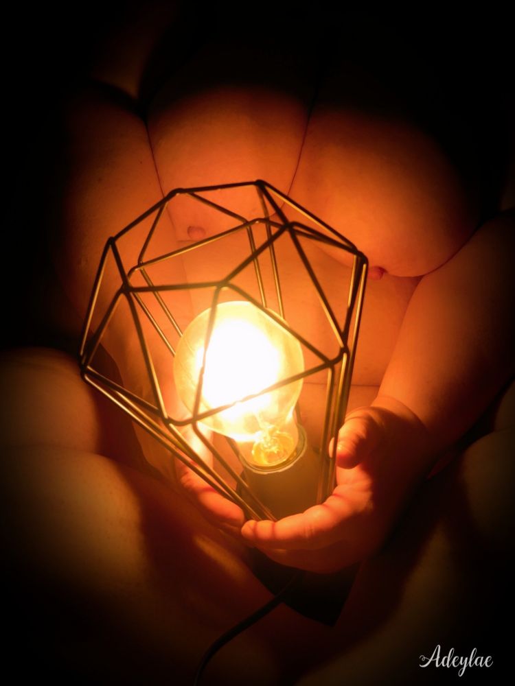 Femme nue assise en tailleur, tenant une lumière entre ses mains. Son corps est plongé dans l’ombre, mais l'éclairage révèle sa poitrine et ses mains, tandis que son visage demeure dans l'obscurité.