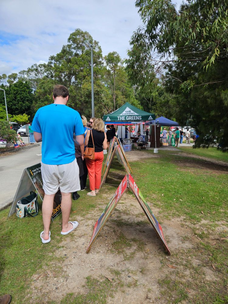 A line of people next to political parties' tents and sandwich board signs at a polling booth