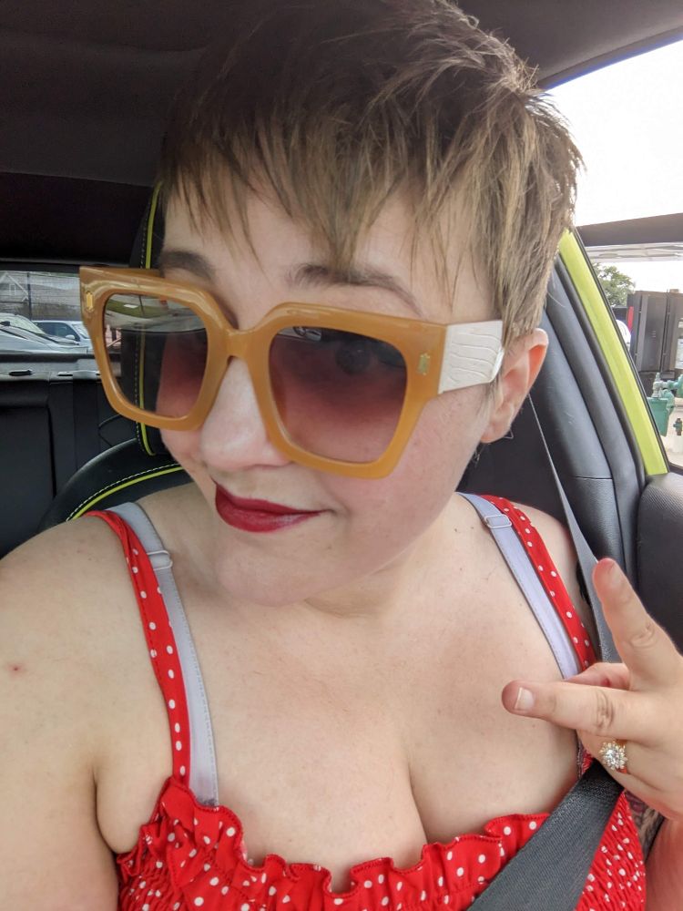 A white woman with a brown pixie cut wears yellow sunglasses, red lipstick, a red polka dot dress, and gives a peace sign.