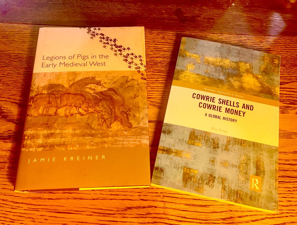 Two books on a wooden table. Left: “Legions of Pigs in the Early Medieval West” by Jamie Kreiner, featuring a medieval illustration of pigs. Right: “Cowrie Shells and Cowrie Money: A Global History” by Bin Yang, with a modern abstract cover design.