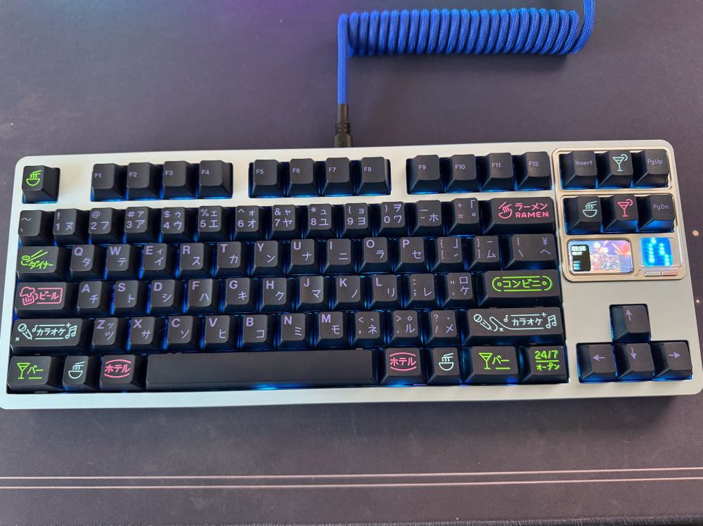 Custom QK80 MK2 mechanical keyboard build with an ice blue aluminum body. The keyboard features an LED screen to show time as well as a fun pixel animation. The keycaps are GMK Night Life, which features Japanese kanji sublegends and fun novelty keycaps representative of night life. There is a coiled blue aviator cable connecting the keyboard to the computer.