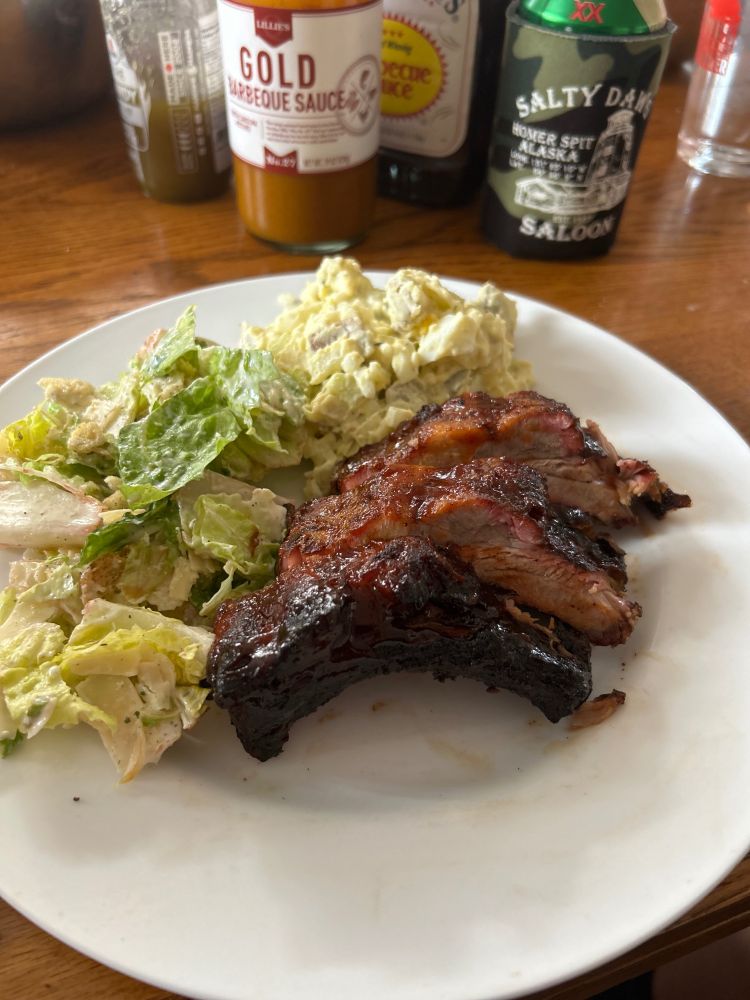 White dinner plate with baby back ribs, potato salad and a Caesar salad on a brown table 