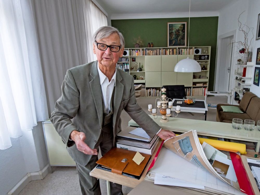 Der ergraute elegante Rudolf Horn im Dreiteiler blättert durch Entwürfe zum Betrachtenden gerichtet mit einer erklärenden Geste. 
