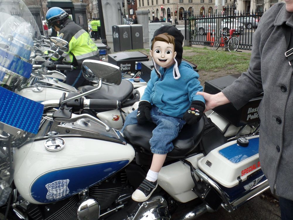 A photo of a toddler in jeans and a blue fleece with a hat and mittens sitting on the seat of a Boston PD police motorcycle while a woman holds him steady. 