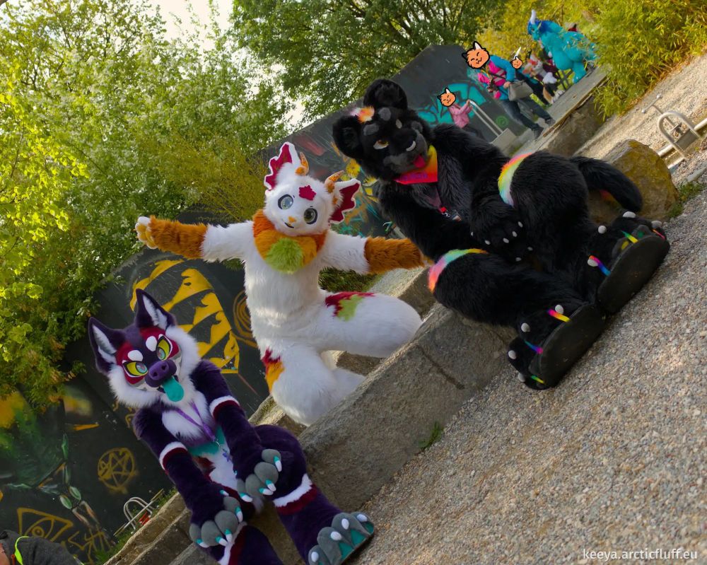 Photo of two fursuiters on a low stone bench in a public park: a purple canine with white markings, yellow eyes, and a long turquoise tongue, and a black jaguar with rainbow stripes. They’re just sitting there with their paws in their laps, looking passive. Unknown to them, a white kemono dragon fursuiter with autumn leaves and orange-green highlights is posing behind them, arms spread out and tilted almost diagonally, while the photographer has also tilted the camera at the same angle, turning the otherwise boring scene into a very cool dynamic photo.