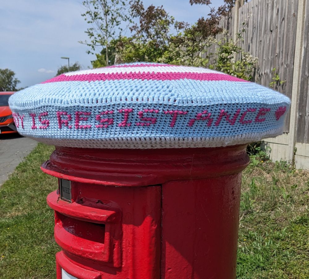 A crochet postbox topper in the trans flag colours with a heart and the message "TRANS JOY IS RESISTANCE" cross-stitched into it (you need to see both pictures to read the full message)