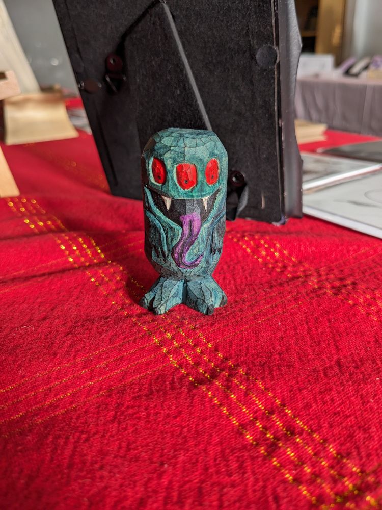 A small, dark green carved wooden monster with a long purple tongue, a wide mouth, two fangs and three red eyes. He's standing on a red tablecloth.