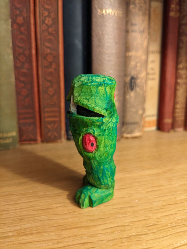 A whittled green monster, seen from the left, with a single red eye in the middle of its thigh, dark green veins and a large tusk visible in its mouth. It has no arms and long chunky legs.
