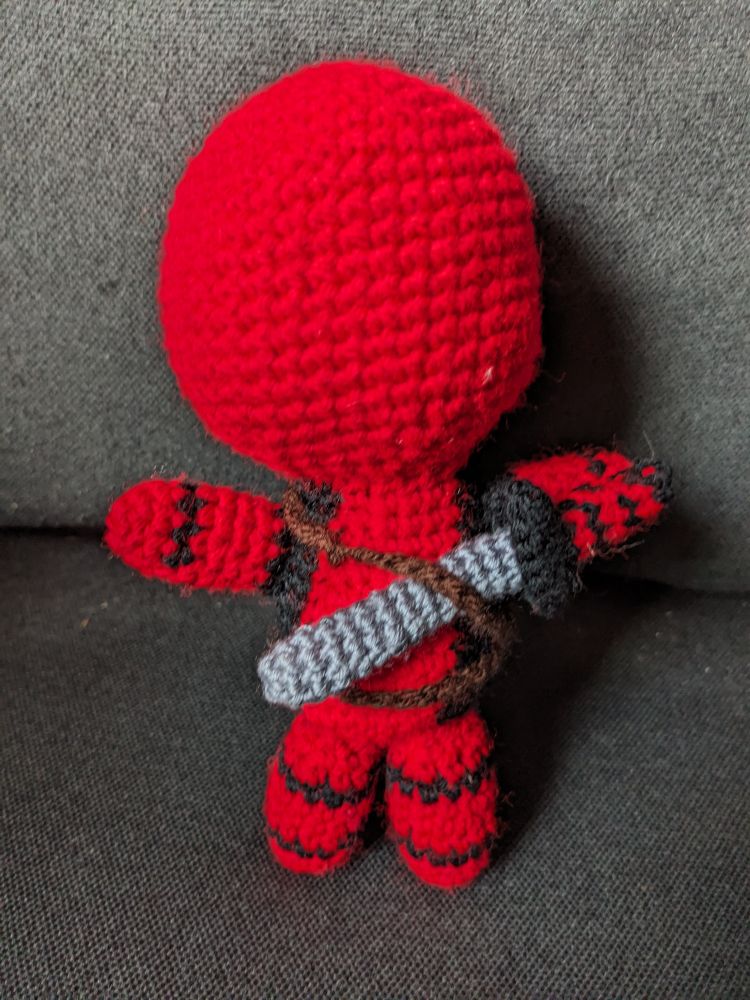 A crochet Deadpool doll seen from the back. He has a crochet sword in a bandolier.