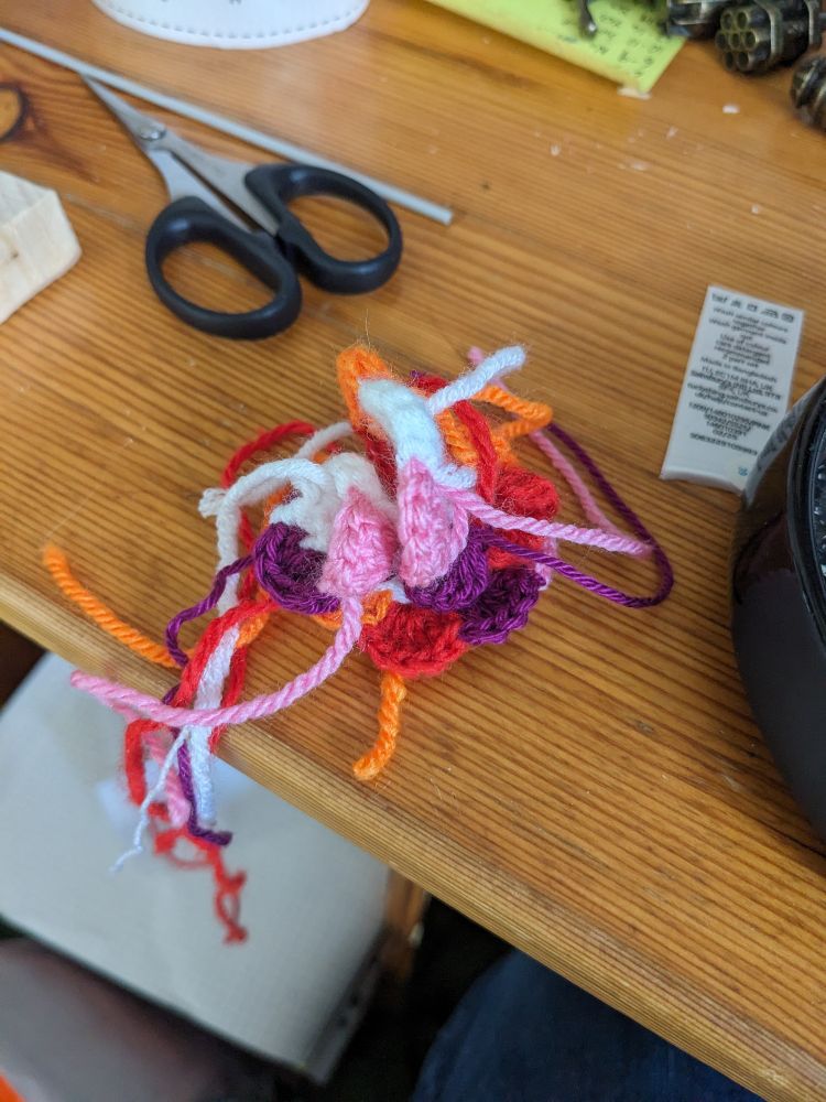 A crochet rose featuring all the colours of one of the lesbian flag designs, with a lot of tangled loose threads.
