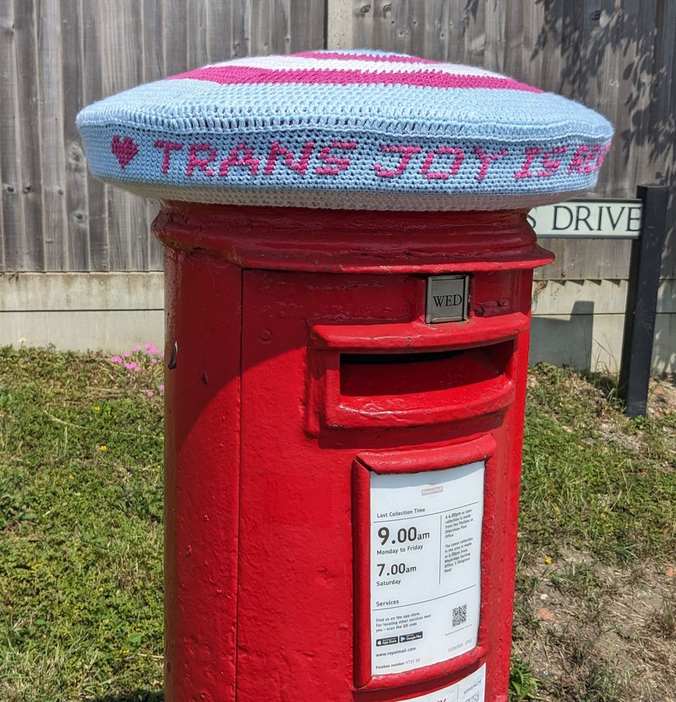 A crochet postbox topper in the trans flag colours with a heart and the message "TRANS JOY IS RESISTANCE" cross-stitched into it (you need to see both pictures to read the full message)