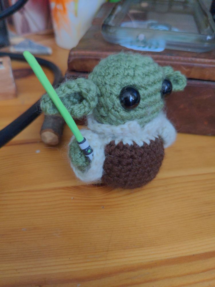 A crochet baby Yoda figure with a painted wooden lightsabre, side view