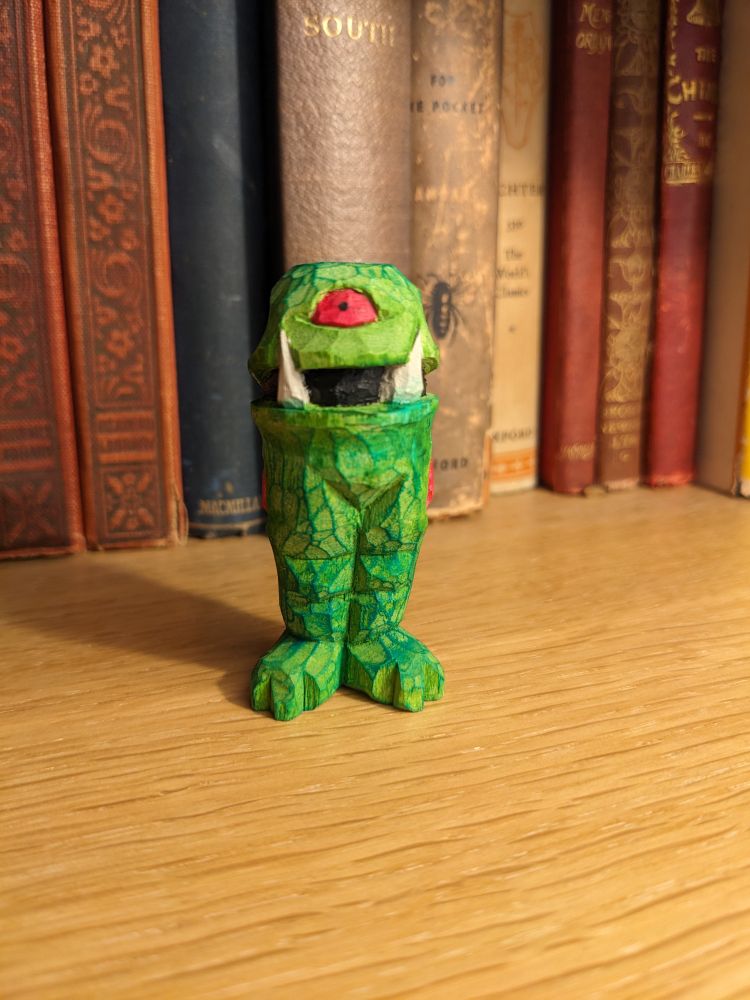 A whittled green monster with a single red eye in its forehead, dark green veins and two large tusks. It has no arms and long chunky legs.