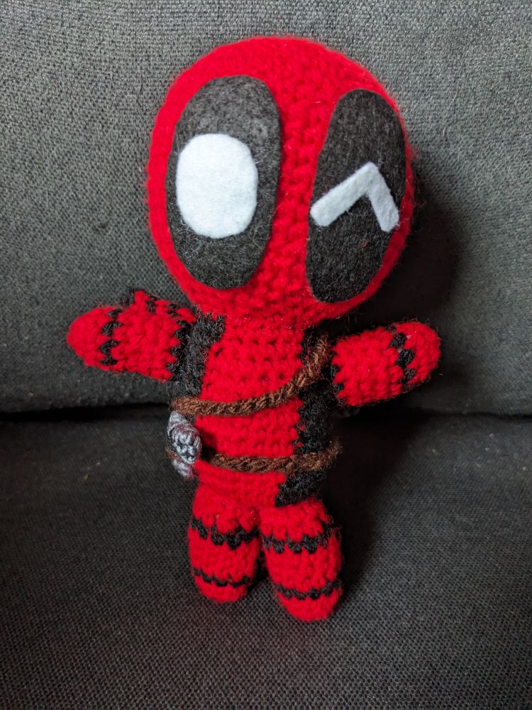 A chibi Deadpool crochet doll with one eye winking and both arms raised as if to give someone a hug. He has a tiny crochet gun too!