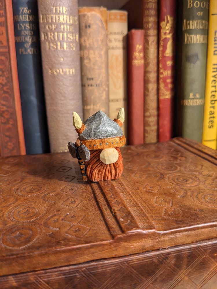 A whittled tomte gnome with a Viking helmet and double-headed axe. He's sitting on a wooden box with old books in the background.