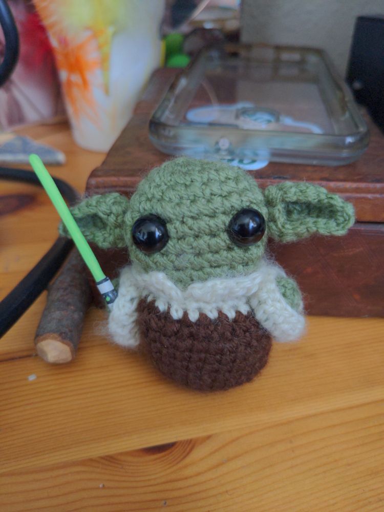 A crochet baby Yoda figure with a painted wooden lightsabre