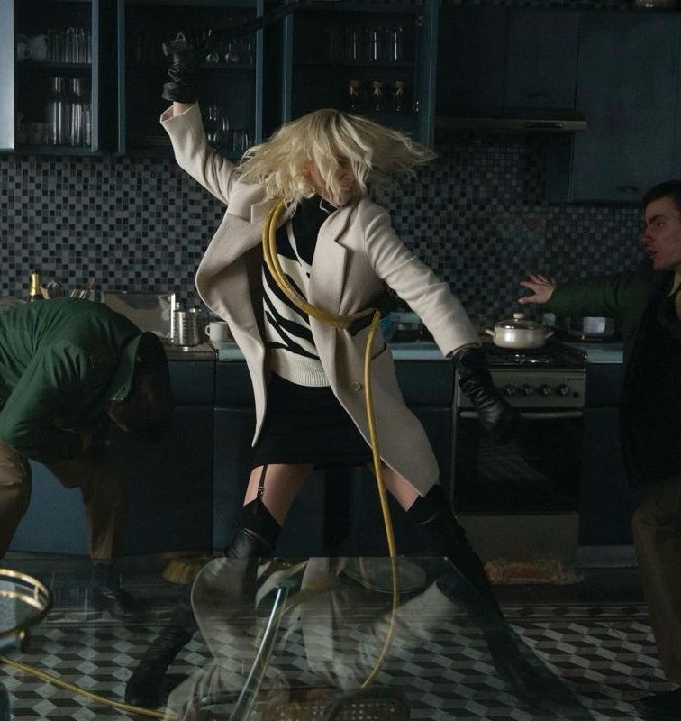 A still from Atomic Blonde. Charlize Theron’s character is in a kitchen, taking a fighting stance with one hand holding a baton, raised and ready to hit the man coming after her. She’s wearing knee high black leather boots, black thigh highs and garter belt, a short black skirt, black and white sweater, and a long white overcoat.