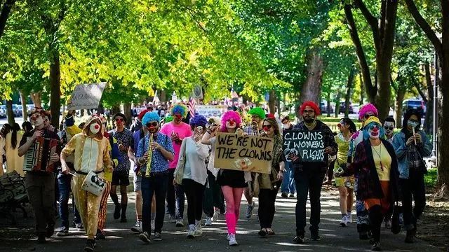 Photograph of a crowd of colorfully dressed clowns marching in a tree-filled park, holding signs saying things like “WE’RE NOT THE CLOWNS” and “CLOWNS 4 WOMENS’ RIGHTS”