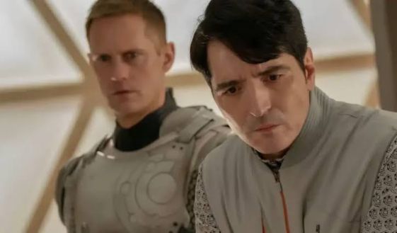 Still frame from the "Murderbot" TV show. SecUnit (played by Alexander Skarsgard, a tall thin young man with short dark blond hair, a high forehead and an angular face) stands to the rear of Gurathin (played by David Dastmalchian, a dark-haired middle-aged man with an angular face and tightly pursed lips). Gurathin is looking down at something in front of him in consternation while SecUnit looks on with a neutral expression.