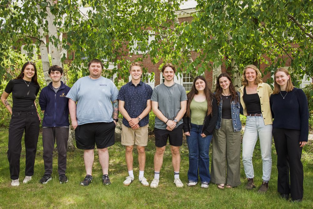 A group photo of the Doyle Fellows, standing under a tree on campus with a brick building in the background. 