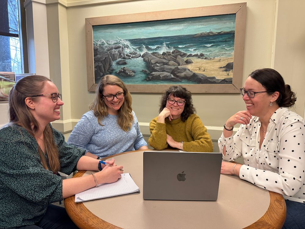 A group of four staff members sit at a table around a laptop, smiling. 