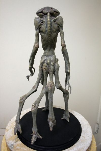 Full body maquette sculpture of an alien with 5 legs and a strange head almost entirely lacking facial features. Back view.