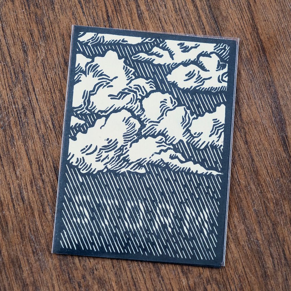 Black and white linoleum block print of a Magic the Gathering storm token card. The rain from the clouds forms the word “storm” in the negative space