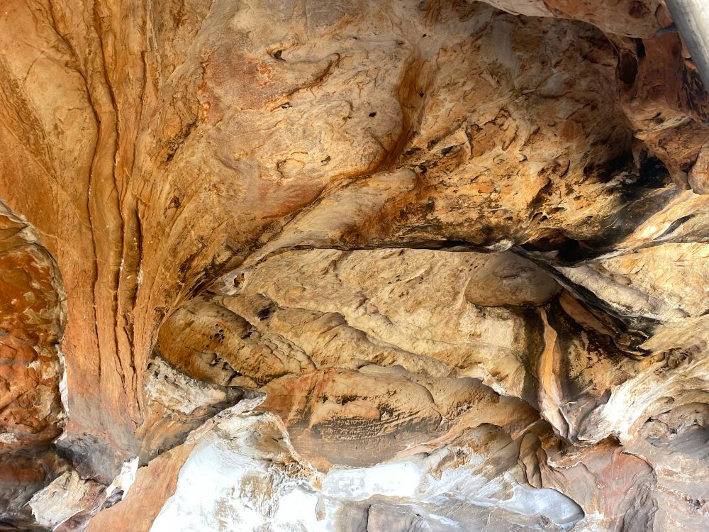 Photograph of ancient aboriginal cave art in a rocky overhang. Red ochre hand/prints, made with the spray method can be seen in various locations, including one way up high in a very inaccessible spot. 