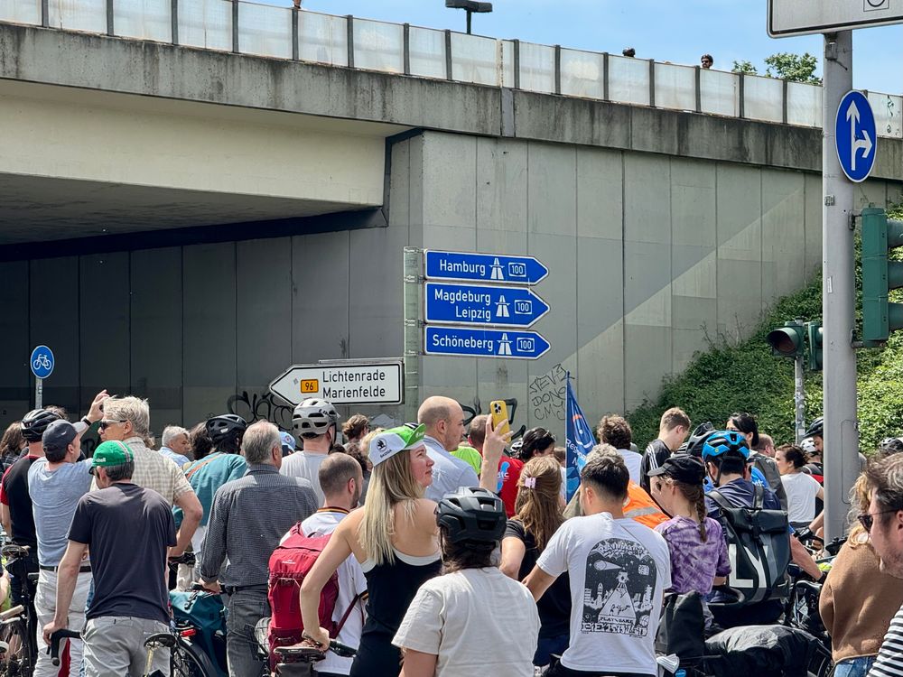 A large group of people, many on bicycles, gathers under an overpass. Directional signs indicate routes to various cities, including Hamburg and Magdeburg. The scene is lively, with individuals holding flags and using their phones.