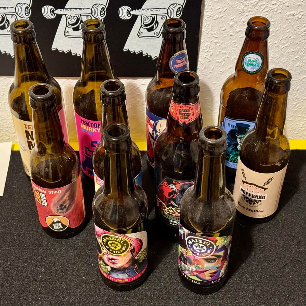 A collection of empty beer bottles with colorful labels displaying various designs and brand names, arranged on a dark surface.