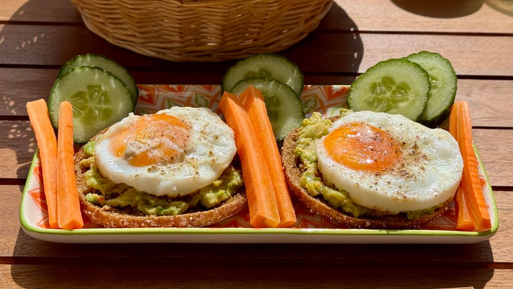 A plate with two toasted slices of bread topped with avocado and sunny-side-up eggs, accompanied by carrot sticks and cucumber slices. The setting appears sunny with a wicker basket in the background.