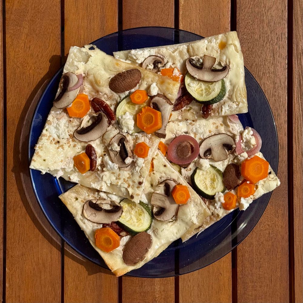 A blue plate holds several slices of flatbread topped with a variety of vegetables, including mushrooms, carrots, zucchini, and red onions, along with crumbled cheese. The plate is placed on a wooden table.