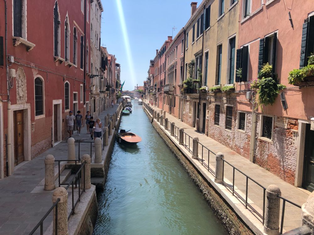 Venedig, ein kleiner Kanal mit rötlichen Häuserzeilen an beiden Seiten. Am Hinmel ein vertikaler weißer Kondensstreifen.