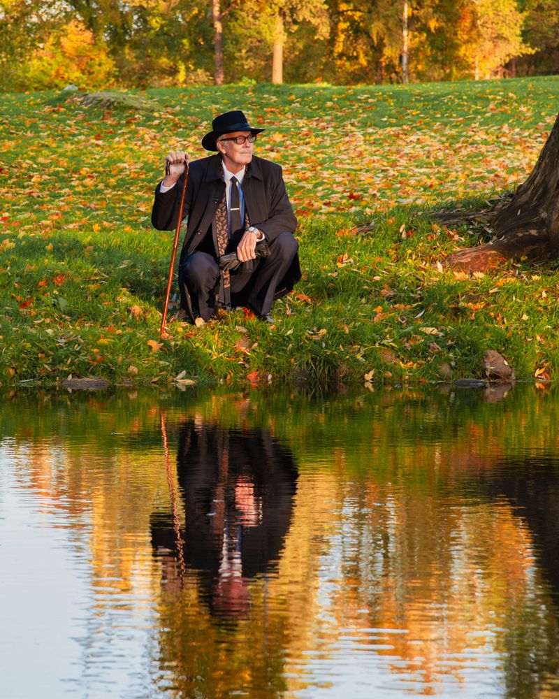 Vanhempi mies vanhanaikaisessa puvussa, hatussa ja silmälaseissa lammen rannalla. Hänen kuvansa heijastuu lammen pinnasta. Taustalla puita syksyn väreissä ja pudonneita lehtiä nurmikolla.