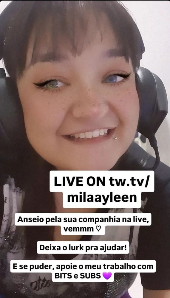 Uma mulher da pele clara com um sorriso bem aberto e mostrando os dentes, a mulher possui cabelos lisos e escuros com franja e um piercing (bolinha) na boca e veste roupa preta com detalhes em braço e usa um headset preto.