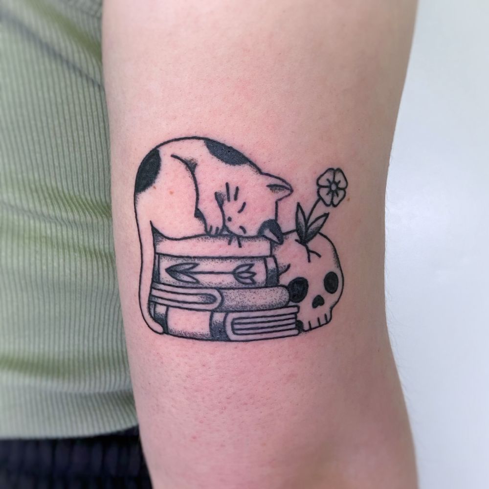 Tattoo of a cat laying on a stack of books with a skull beside