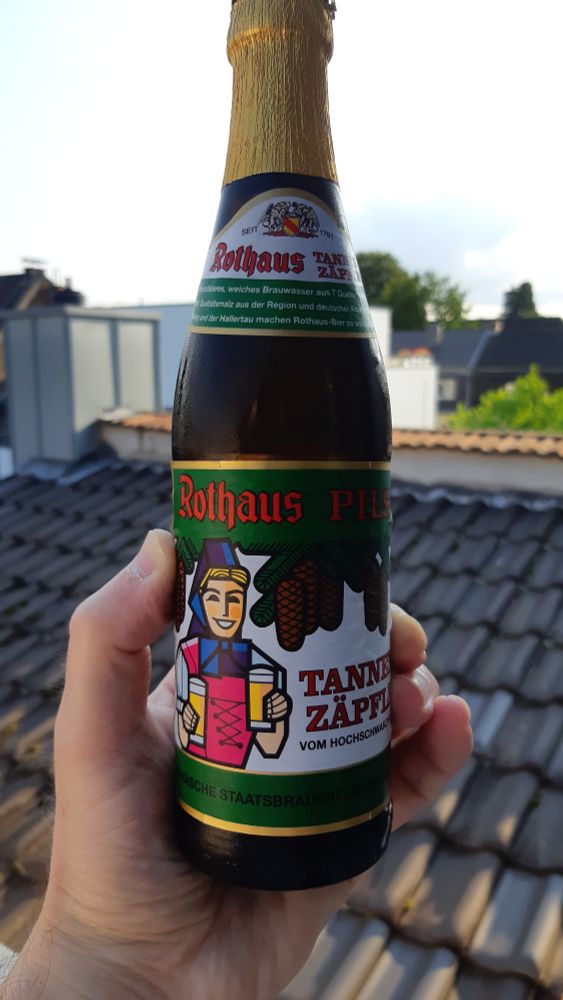 Foto einer Hand, die eine Bierflasche der Marke Tannenzäpfle, Rothaus Pils hält. Im Hintergrund sind Dachpfannen umd unscharf ändere Dächer zu erkennen, zusätzlich fast blauer Himmel in der Abendsonne. #AufMicha