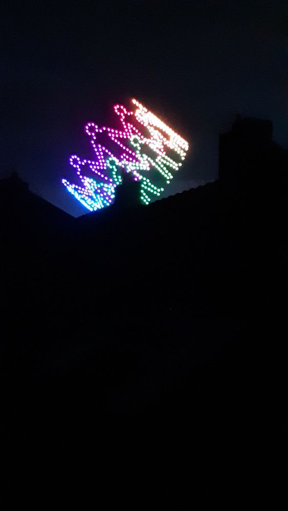 Dunkler Nachthimmel. Zu sehen ist eine Drohnenshow. Ein Kreis von Menschen halten sich an den Händen, das Motiv ist in Regenbogen-Farben gehalten.