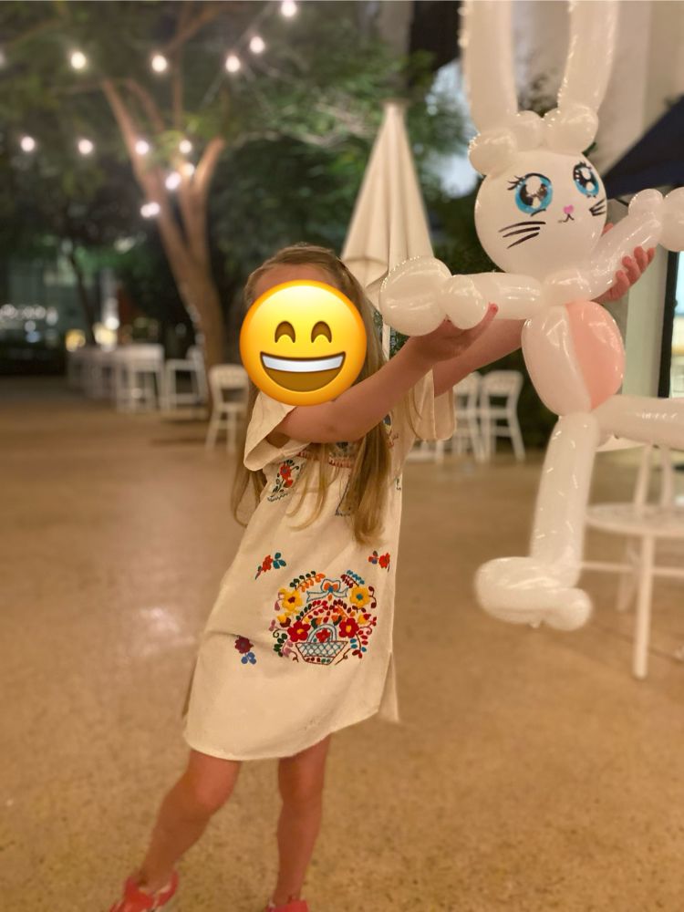 Girl wearing a white linen dress with multicoloured floral embroidery, joyously displaying a top notch bunny balloon animal. 