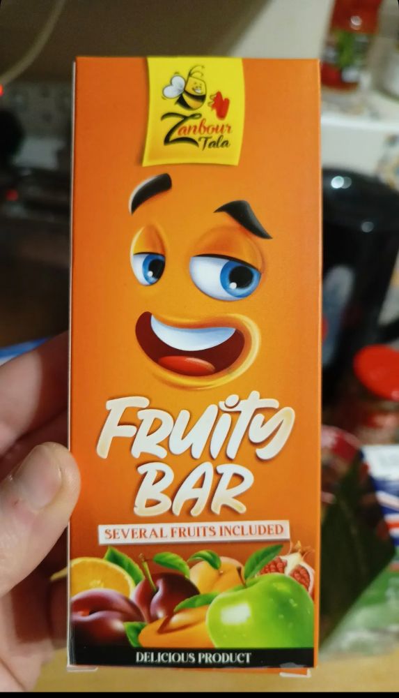 Product packaging - "(zanbour taka)
 Fruity Bar
Several fruits included"

Weird orange background with a disembodied face on it, like if we were very zoomed in on an orange with a tiny face