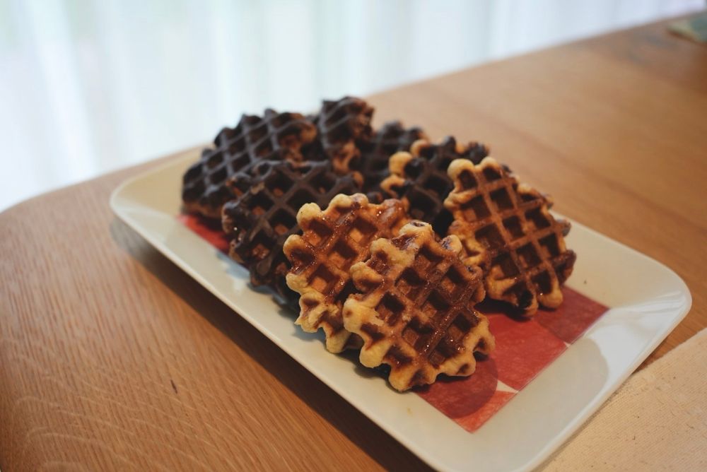 チョコレートがかかったワッフルとプレーンなワッフルが複数四角い皿に乗せられている様子。皿は木製のテーブルに乗っている。