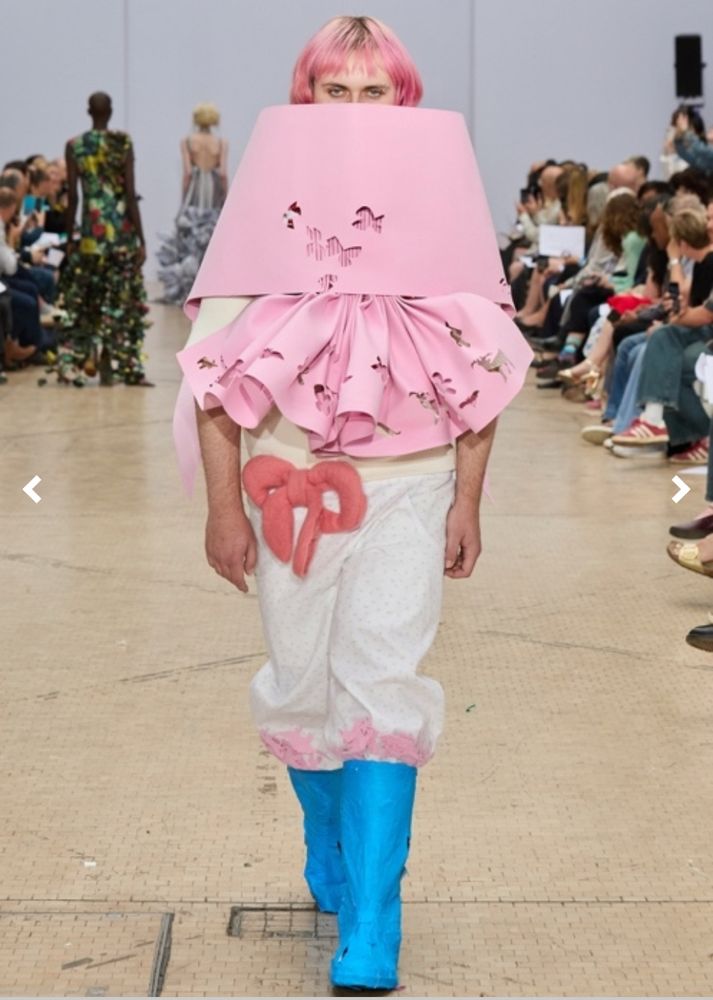 Human walks a fashion catwalk wearing very blue rubber knee-high boots, white pants with pink edging and a light pink giant lampshade that serves as both a crop top and turtle neck. 