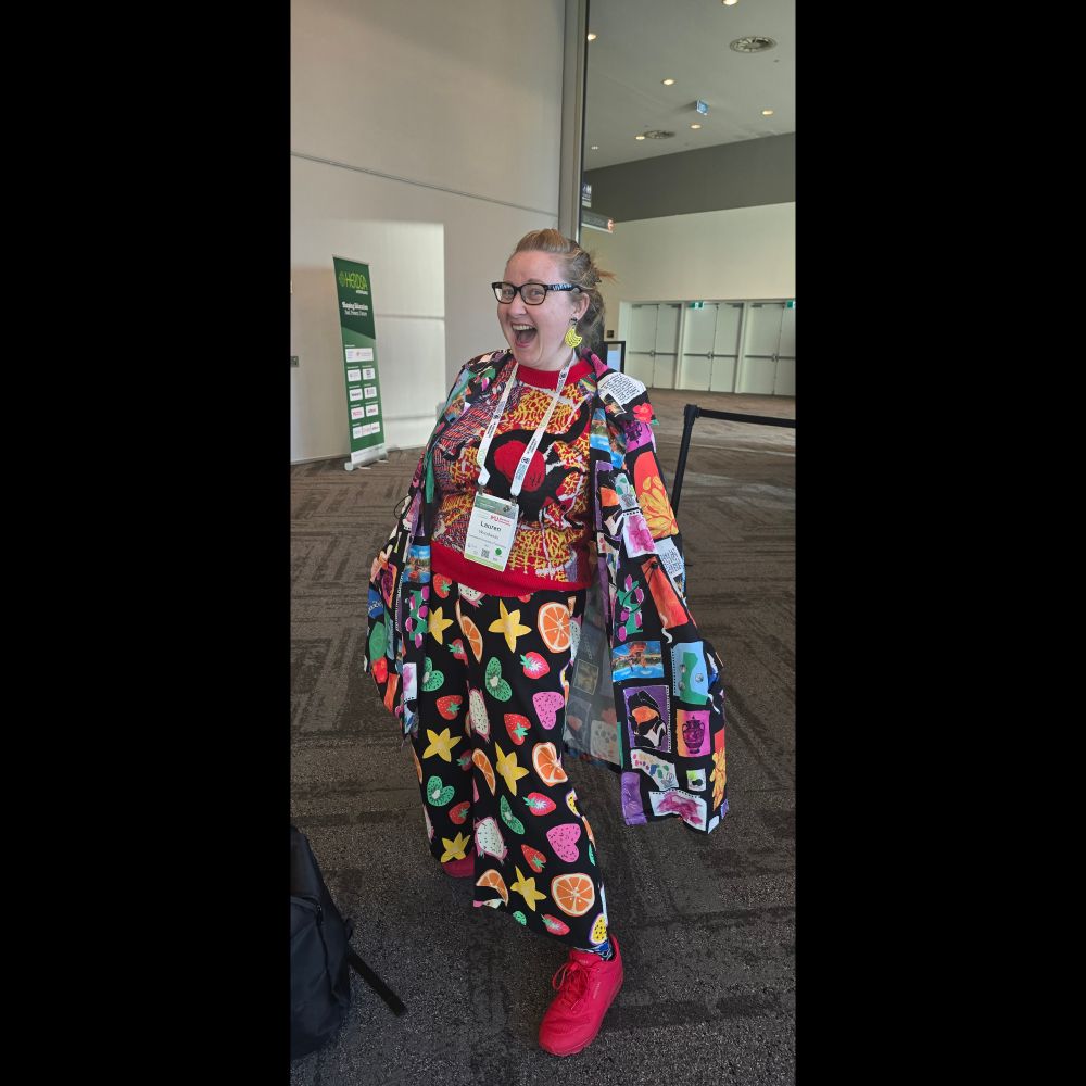 Lauren leaning back holding out her garishly coloured and patterned jacket,  which clashes with her bright coloured fruit covered pants and luminescent shoes. 