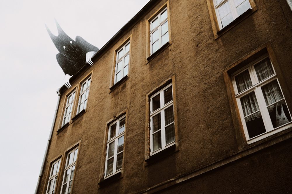 Eine Hausfassade in goldbeige. Über das Haus beugt sich eine schwarze Kreatur, die in eines der weißen Fenster schaut, in dem eine kleine weiße Kreatur unter einer Gardine hervorlugt