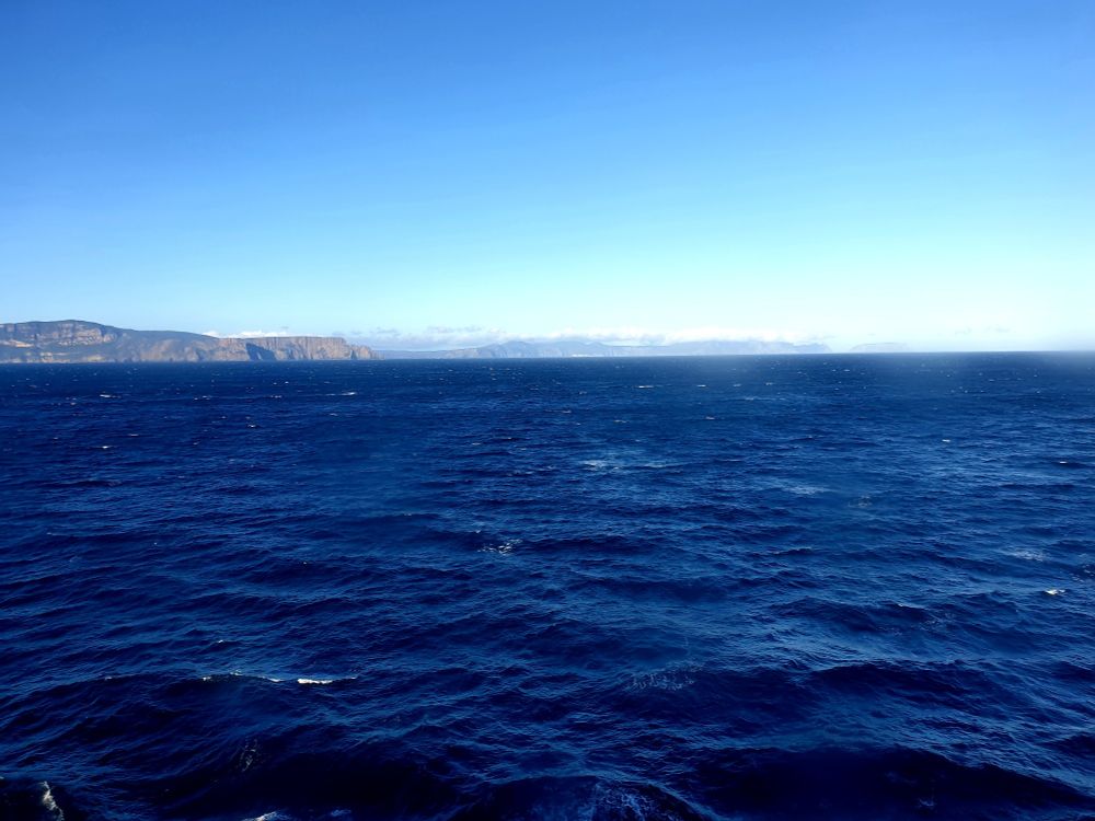 A photo where the frame is mostly filled with the sea, as captured from deck 9 of the Disney Wonder. In the distance land can be seen. Above that is a beautiful blue sky.