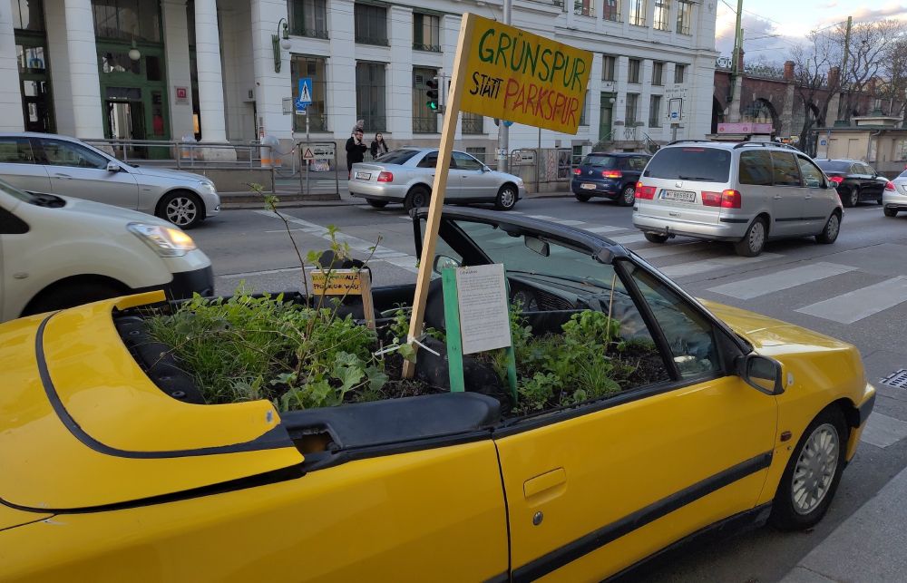 Das Foto zeigt ein gelbes Cabrio mit offenem Verdeck von der Seite. Es ist mit Erde und Pflanzen gefüllt, auf einem darin befestigten Schild steht handgeschrieben der Slogan „Grünspur statt Parkspur“. Das Cabrio steht am rechten Rand einer mehrspurigen Straße. Daneben und davor sind fahrende Autos zu sehen, im Hintergrund eine U-Bahn-Station und zwei Erwachsene mit einem kleinen Kind, die darauf warten, die Straße überqueren zu können.