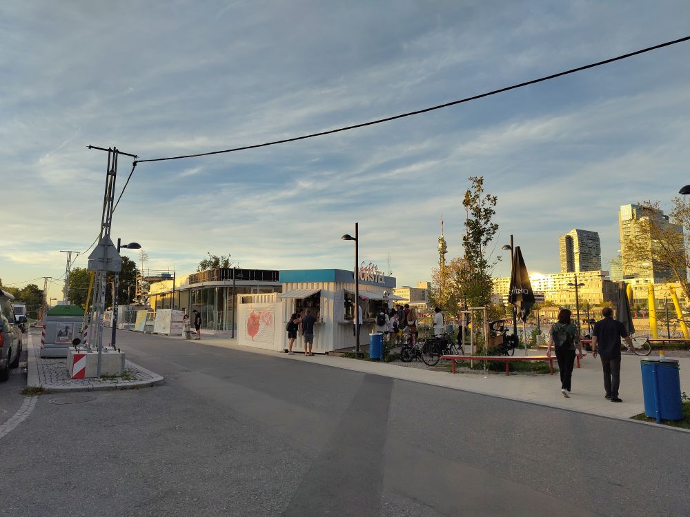 Das Foto zeigt eine asphaltierte Straße mit einem Würstelstand und einem großen, flachen Gebäude links im Hintergrund. 