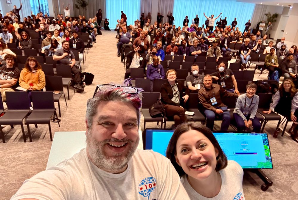 Here is the alt text for tA selfie of two people in the foreground, both wearing light-colored t-shirts with a circular logo featuring gears and a red star. The person on the left has short, graying hair with streaks of color and glasses resting on their head, while the person on the right has shoulder-length dark hair. They are smiling at the camera. Behind them, a large audience of diverse individuals is seated in rows of chairs, many of them smiling, waving, or making gestures towards the camera. Some attendees are wearing lanyards with badges. The room has a modern conference setting with a neutral-colored carpet and bright lighting. Two large screens displaying a presentation slide are visible in front of the speakers.